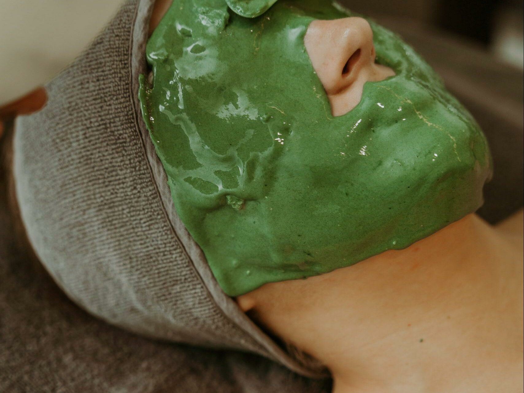 A person with their head wrapped in a gray towel is lying down and receiving a green facial mask. A hand is seen applying the mask with a spatula. The person's eyes are closed, and they appear to be relaxed. The background is blurred and neutral.