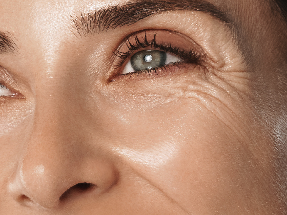 Blog Images Swap-07 Close-up of a smiling person's eye and surrounding area, showing fine lines and a slightly furrowed brow. The skin has a natural glow, with light reflecting off the cheek and eyelid. Brown eyebrows and eyelashes frame the blue eye.