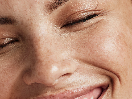 Blog Images Swap-09 Close-up of a person's face with closed eyes and a smile, highlighting clear skin with freckles, reflecting the benefits of green chemistry in skincare.