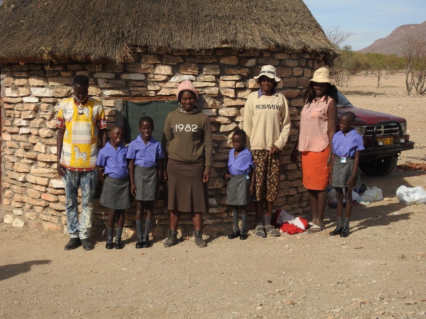 A group of people, including four children and four adults, stands in front of a stone hut with a thatched roof. The children are wearing purple school uniforms, and the adults are wearing casual clothing. The setting appears to be rural with a vehicle in the background.