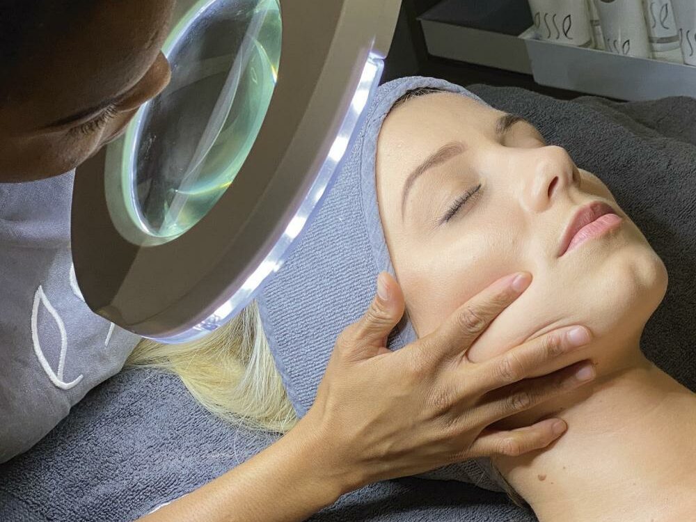 A person is receiving a facial treatment at a spa. Another individual is gently holding their face and examining it under a large magnifying lamp with built-in lights. Skincare products are visible in the background.