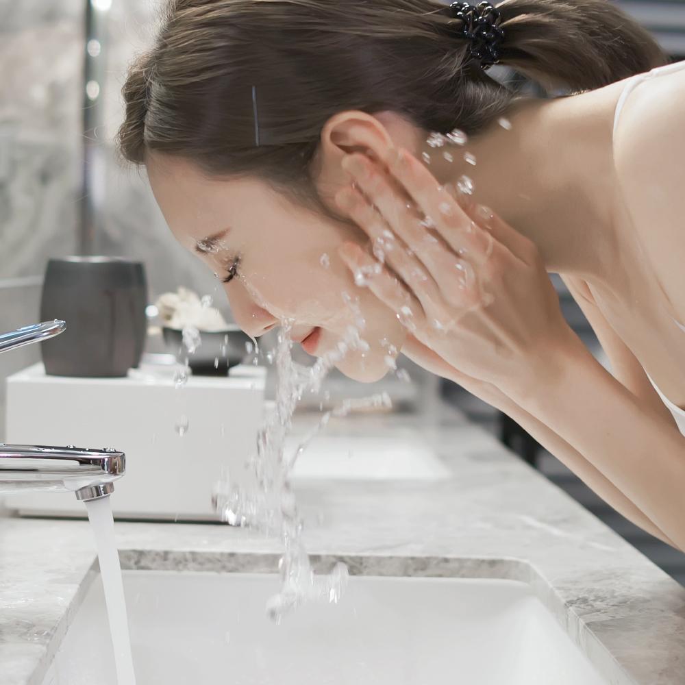 A woman with her hair tied back washes her face over a bathroom sink. Water splashes around her as she leans forward with her hands cupping the water from the running faucet. The bathroom has a modern design with a marble countertop and silver fixtures.
