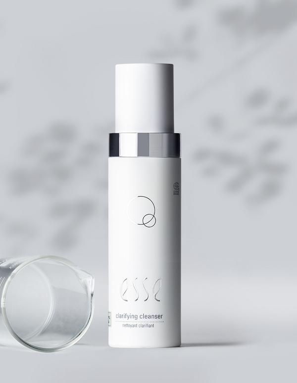 A white bottle of Esse Clarifying Cleanser with a metallic cap stands upright against a light gray background featuring subtle leaf patterns. A clear beaker lies on its side next to the minimalist-designed bottle, which features a circular logo.