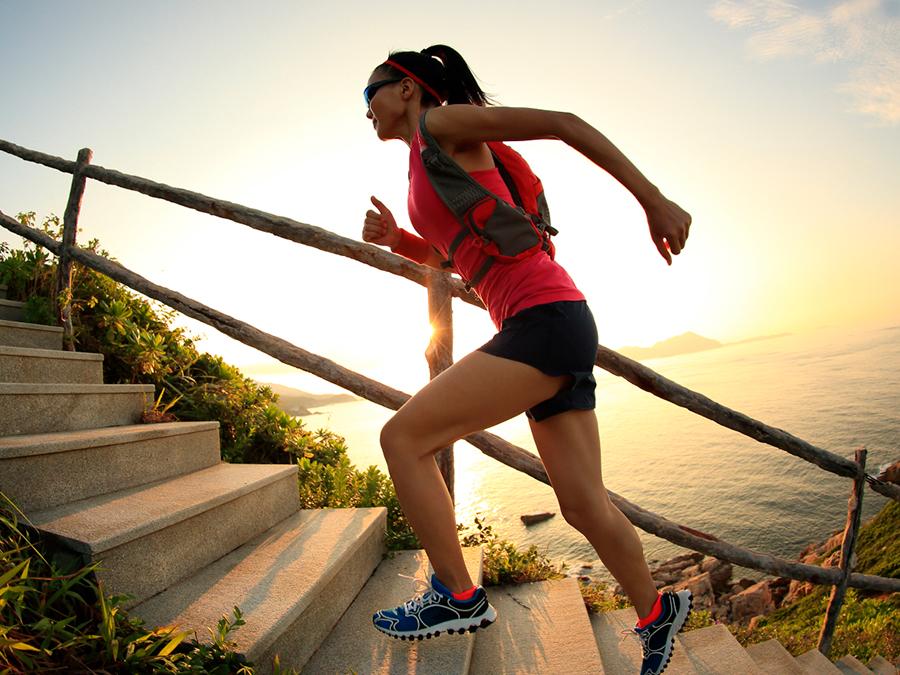 A woman in athletic gear runs up stone steps on a coastal path during sunrise. She wears a red tank top, black shorts, sunglasses, and a hydration pack. The sun illuminates the horizon over the ocean, with lush greenery along the steps.