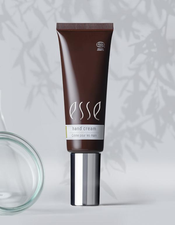A brown tube marked "Esse Hand Cream" with a silver cap stands upright against a light background adorned with subtle leaf shadows. The label features text in both English and French. On the left side, a transparent glass object is partially visible.