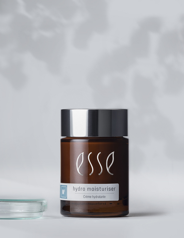 A brown jar with a metallic lid, labeled "Esse Hydro Moisturiser" in white text, stands against a soft, blurry background. The jar features a small blue square with the letter "M" on it and additional text "Crème Hydratante" beneath the primary label. A partially visible transparent glass item is seen on the left side.