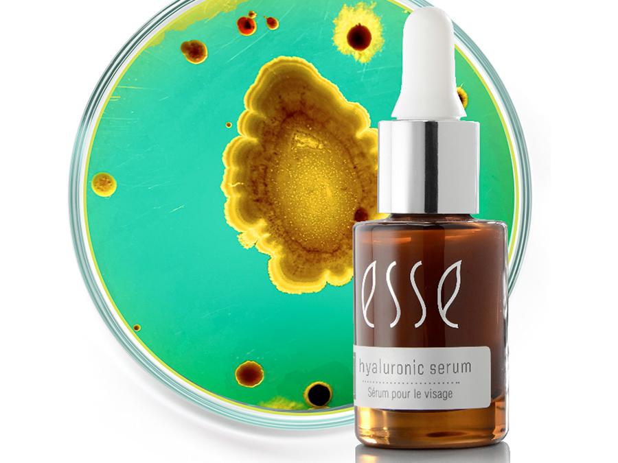 Moisture-andageing—how-the-HA-works A small brown glass bottle labeled "Esse Hyaluronic Serum" with a white dropper cap is positioned in front of a green and yellow petri dish with various circular patterns, suggesting a scientific or skincare-related context.