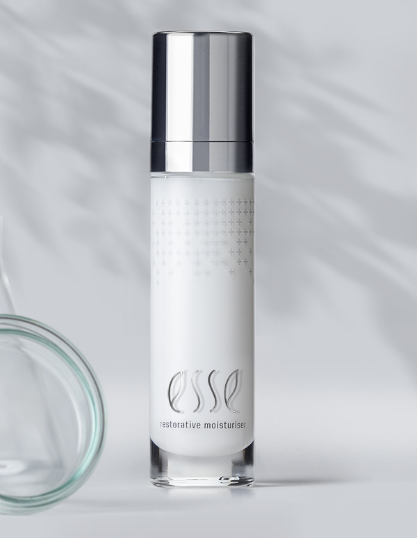 A sleek, silver-capped bottle of Esse Restorative Moisturiser stands against a light grey background. The bottle boasts a minimalist design with subtle text and a faint dot pattern. Shadows from leaves are cast on the background, adding a natural touch.