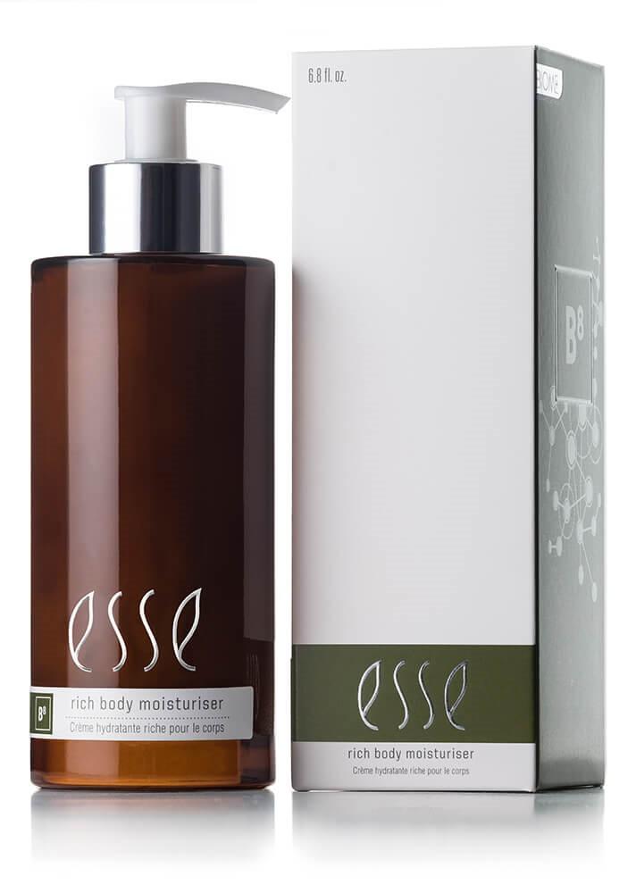 A bottle of Esse Rich Body Moisturiser, made from dark amber glass and equipped with a pump dispenser, is positioned beside its packaging. The predominantly white box with green accents showcases detailed product information and branding. Both the bottle and the box prominently feature the Esse logo.