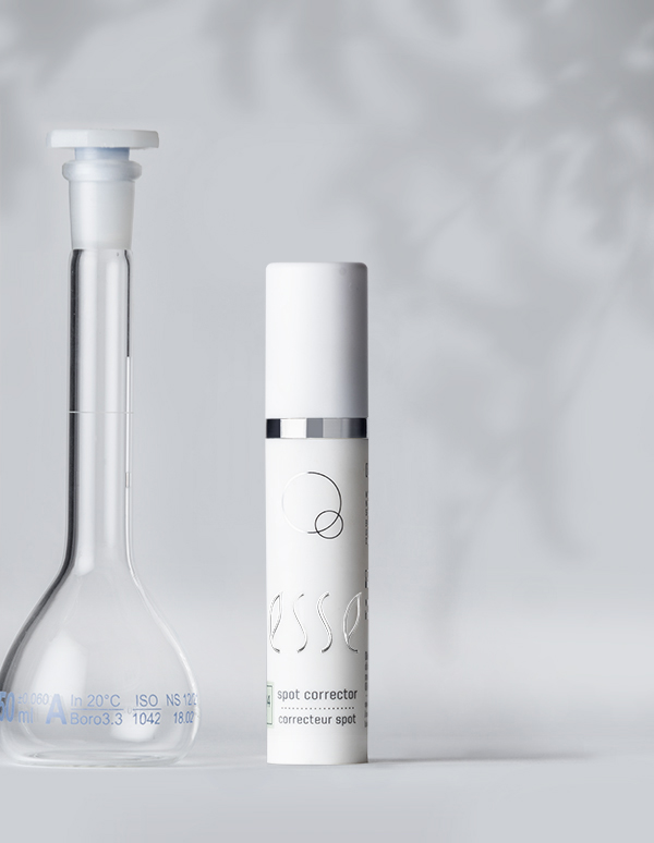 A clear lab flask with a white stopper stands beside a sleek white cylindrical bottle labeled "Esse Spot Corrector" on a light gray background. The minimalist design emphasizes clarity and purity, suggesting a focus on skincare or cosmetic products.