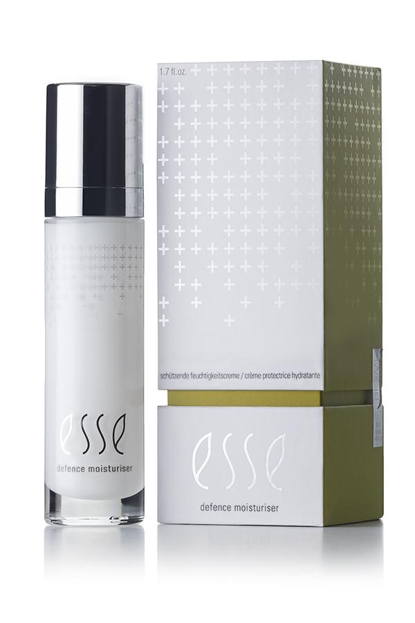 A sleek white and green box with silver accents is positioned beside a cylindrical bottle featuring a shiny metallic cap. The product, labeled "Esse Defence Moisturiser," showcases a minimalist design highlighted by a white background and subtle cross patterns.