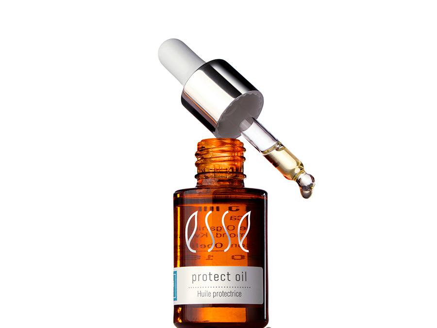 Announcing an addition to the Esse Sensitive line A brown glass bottle of protect oil with a label reading "Huile protectrice." The bottle is topped with a silver and white dropper, which is raised above the bottle, dispensing a drop of oil. The background is plain white.