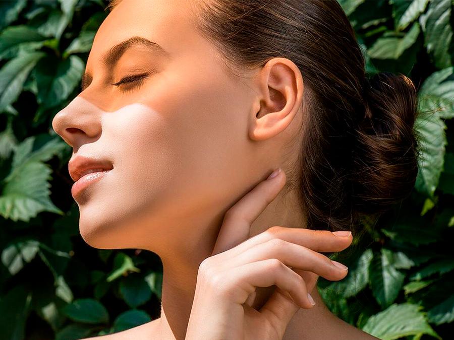 Learn to love your microbes A person with smooth skin and closed eyes stands against a background of green leaves. They have their hair tied back in a bun and are lightly touching the side of their neck with their hand. Sunlight casts a warm glow on their face.