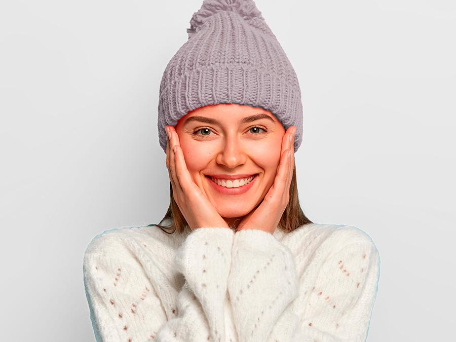 A person wearing a lilac knitted beanie and a white sweater is smiling warmly with their hands resting on their cheeks. The background is plain white.
