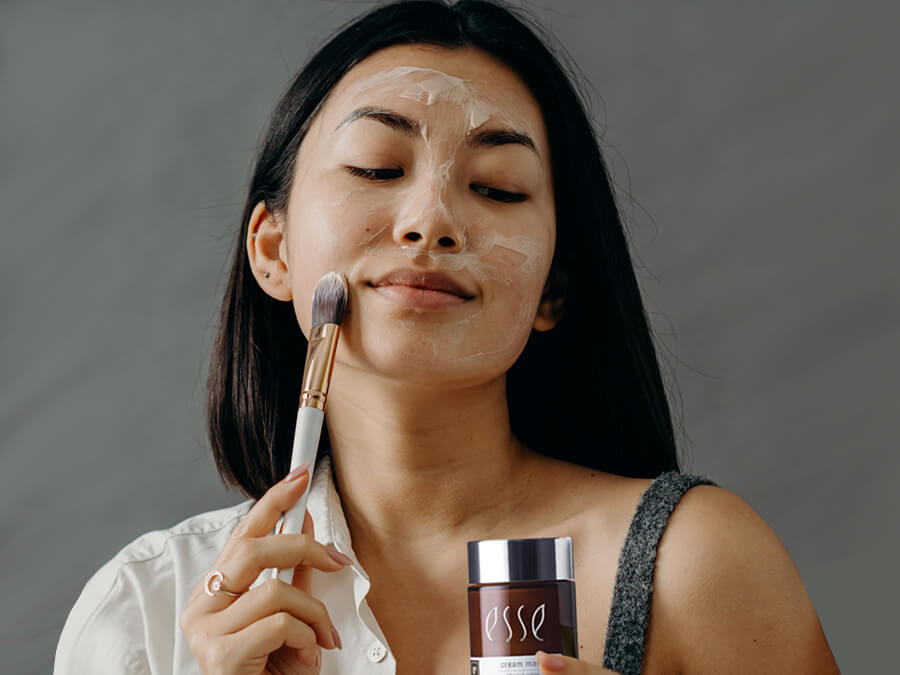 A woman is applying a facial mask using a brush. She is holding a container labeled "Esse" in her left hand. The background is neutral and she appears relaxed.