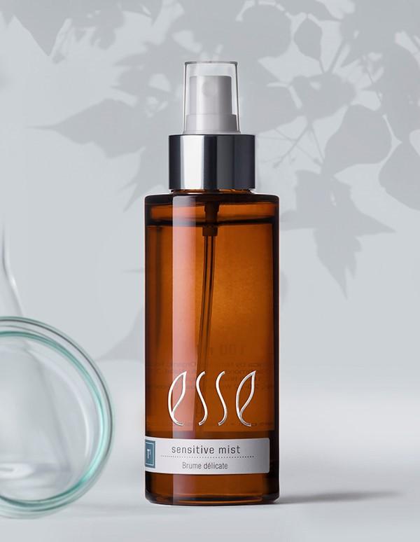 A brown spray bottle labeled "Esse Biome Mist" with a metallic silver pump and white cap. The background contains leafy shadows, giving a natural and serene ambiance. A clear glass container is partially visible on the left side of the image.