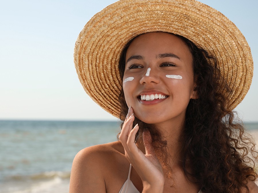 A smiling woman with curly hair wearing a straw hat, standing at a beach with the ocean in the background. She has sunscreen applied on her cheeks and is touching her face gently.