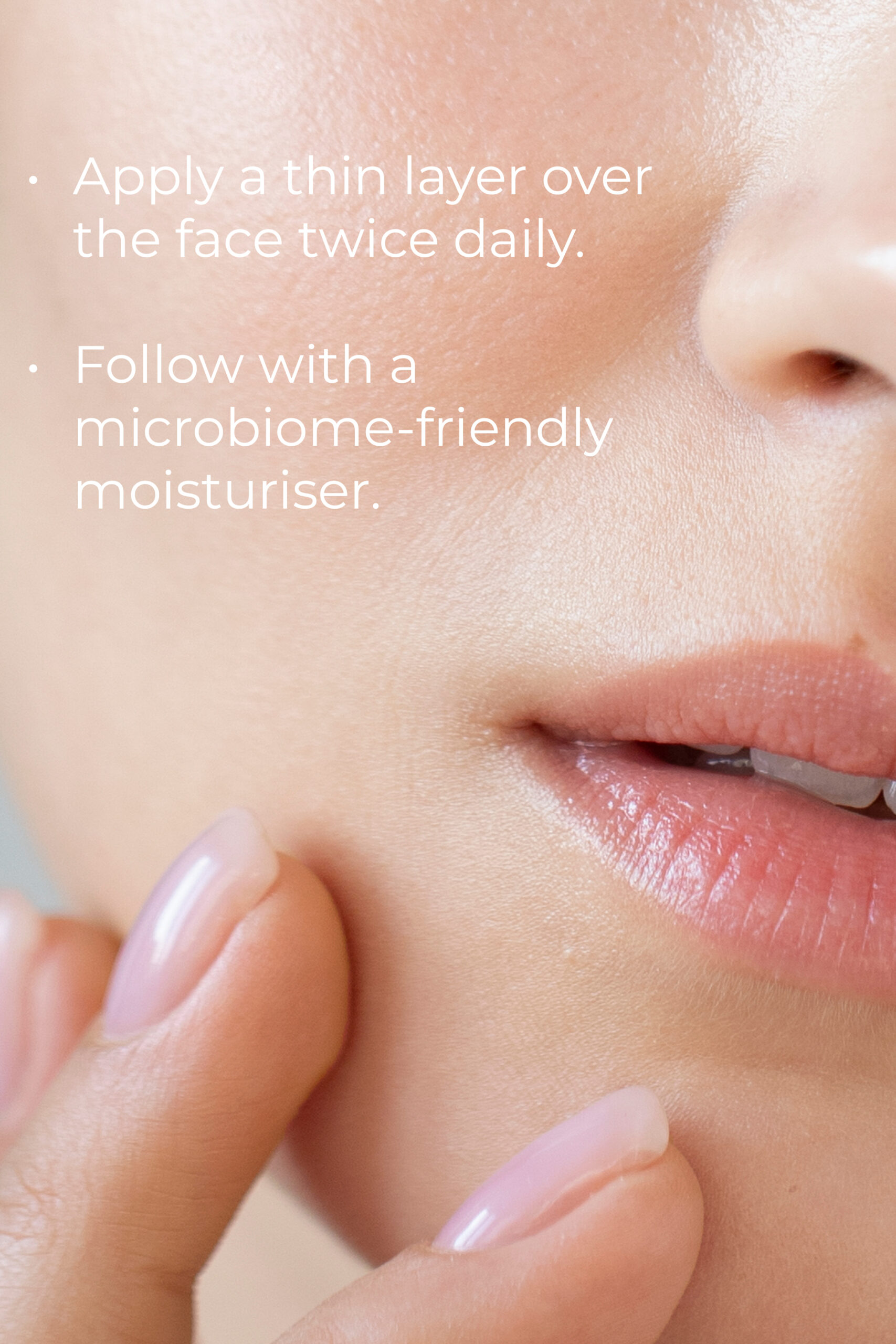 Close-up of a person's face, highlighting the lips and lower cheek. The person has smooth, clear skin and is touching their cheek with their fingers. Instructions on the image read: "Apply a thin layer over the face twice daily. Follow with a microbiome-friendly moisturiser.