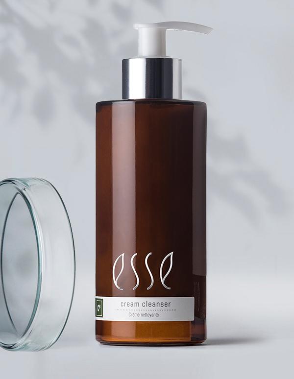 A brown bottle of Esse cream cleanser with a silver pump top, positioned next to a clear glass lid. The label on the bottle includes the product name and some information in smaller text. A shadow of a plant is visible in the background on a light surface.