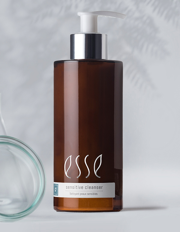 A brown bottle of Esse Sensitive Cleanser with a silver pump sits in front of a blurred background. The white label has the branding "Esse" and the text "Sensitive Cleanser - Nettoyant peaux sensibles." A glass object is partially visible on the left side.