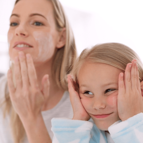 A woman is applying skincare cream to her face while smiling. Next to her, a young girl with long blonde hair is mimicking the action, touching her cheeks with a playful smile. Both are wearing casual, light-colored clothing, and the background is softly blurred.
