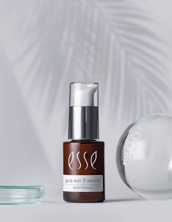 The Esse Pro-Sun D Serum bottle, featuring a brown body with a white and silver pump dispenser, is set against a light background with the soft shadow of a palm leaf. Next to the bottle are a translucent jar cover and a glass surface.