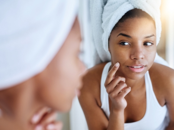 1 A woman with a towel wrapped around her hair looks at herself in the mirror, gently touching her cheek with one hand. She appears to be inspecting her skin. The setting suggests a bathroom, and she is wearing a white tank top.